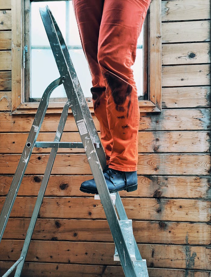 A person stands on a ladder in orange pants, working on a wooden wall beside a window.